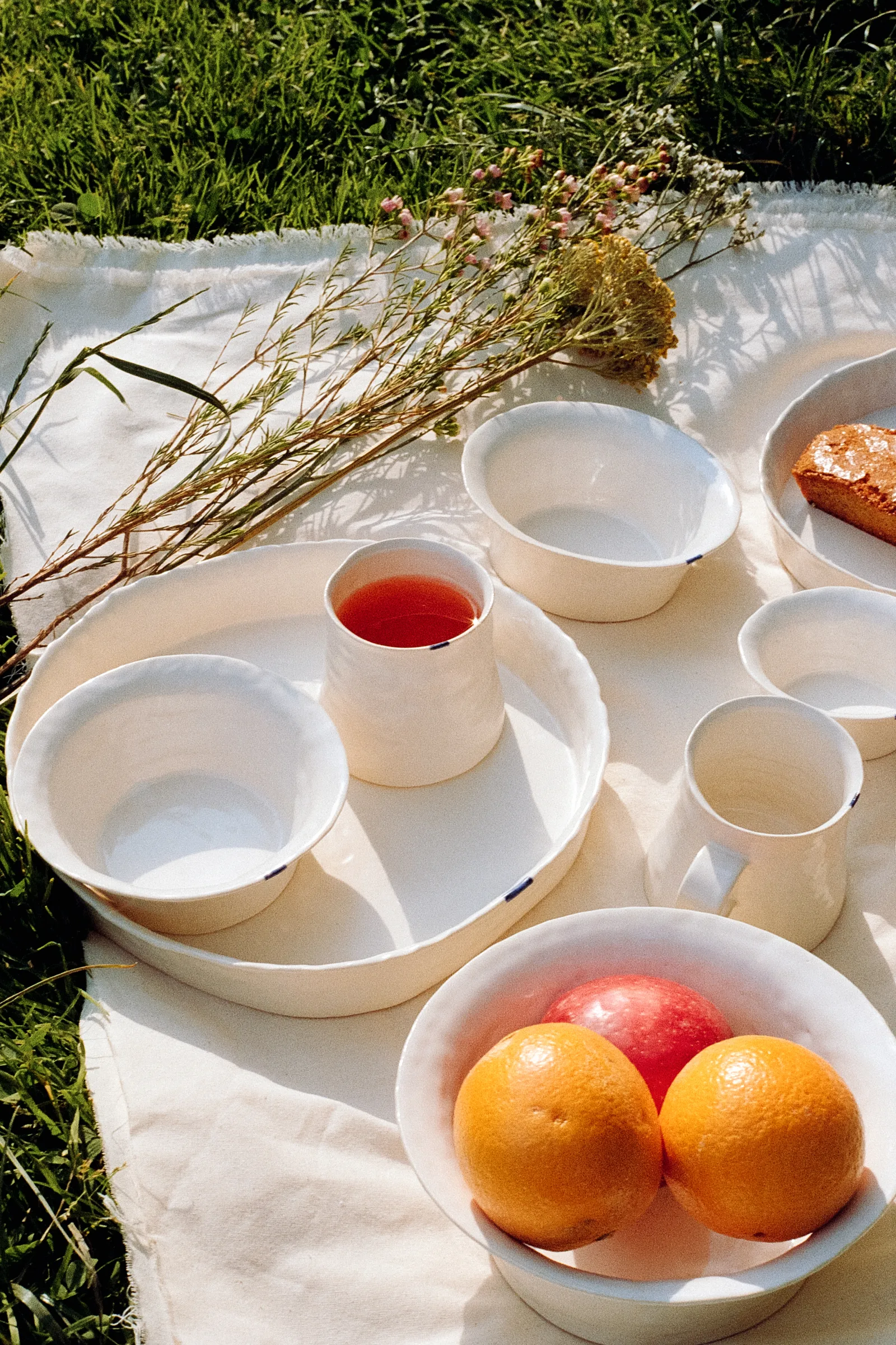 White ceramics, fruit, tea, and flowers arranged on a picnic blanket